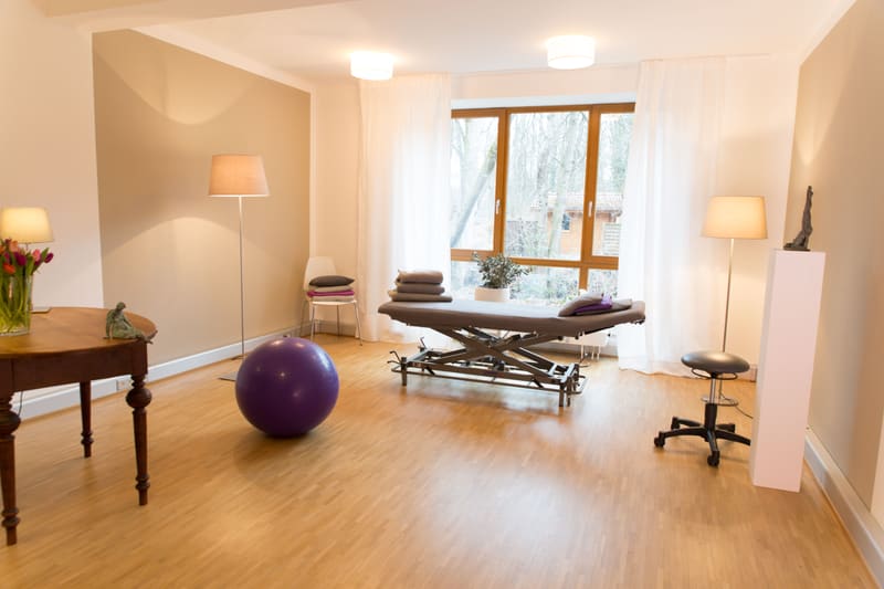 Behandlungsraum mit Behandlungsliege, Gymnastikball und großem Fenster zum Garten